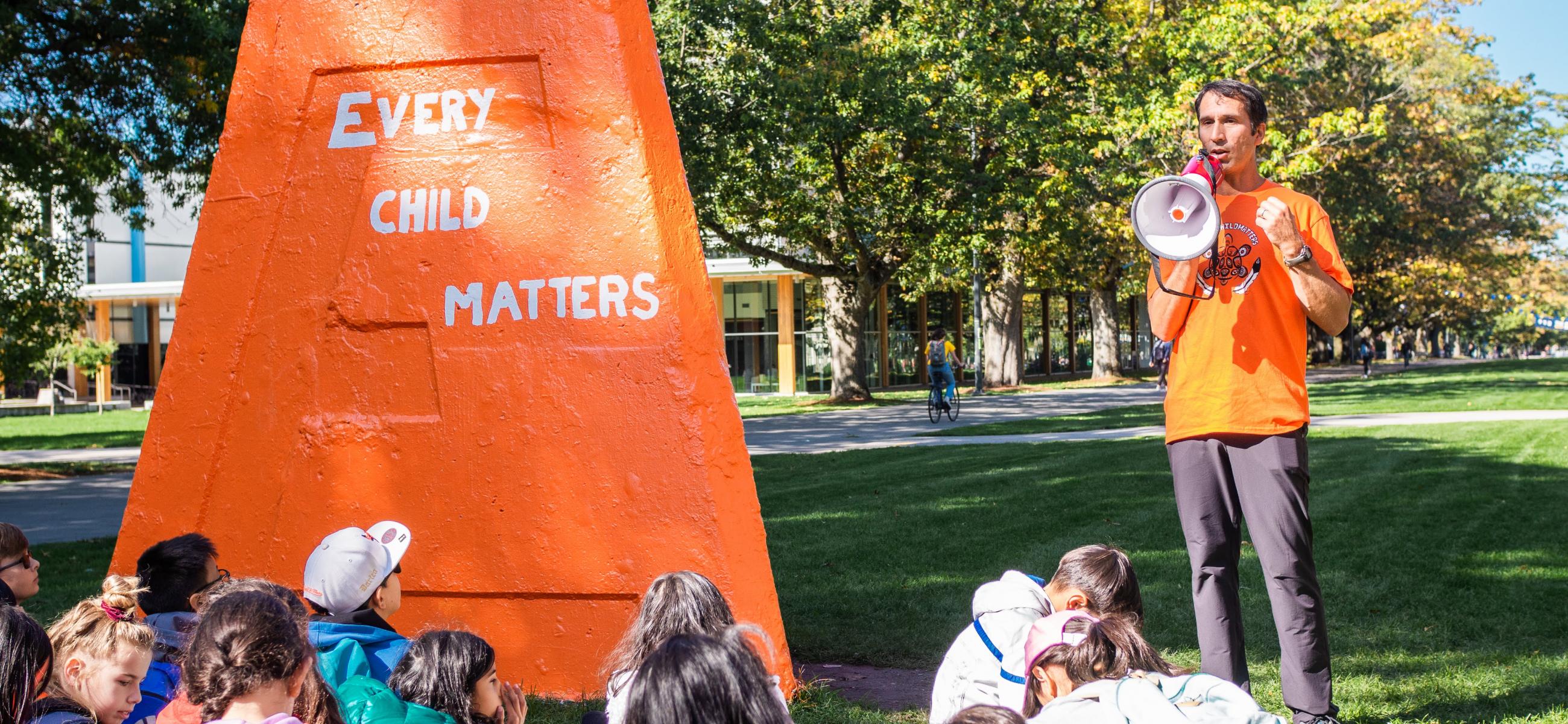 Danilo Caron in an orange shirt, speaking next to the UBC Engineering Cairn painted orange with "Every Child Matters" on it.