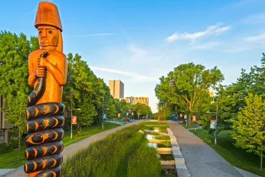 A tall totem pole featuring a human figure holding a serpant that wraps around them, stands at the entry to a long tree-lined boulevard.