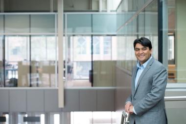Dr. Rehan Sadiq smiles at the camera, casually standing at the top of a set of stairs inside a modern campus building.
