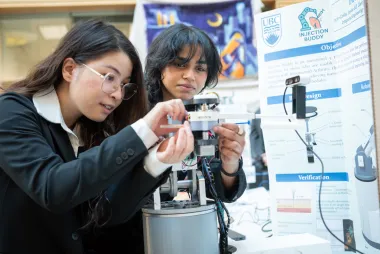 Two students examine a robotic device in front of a poster explaining the project. 
