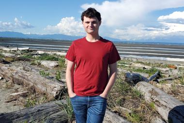 UBC Mechanical Engineering stuent Josh Heieis smiles for the camera in a red shirt, on the shoreline with the tide out in the distance behind him.