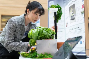 Student appears to be working on a project related to sustainable technology, possibly involving plant growth or environmental innovation, as they are examining a plant-growing setup, with research posters in the background explaining the design and analysis process