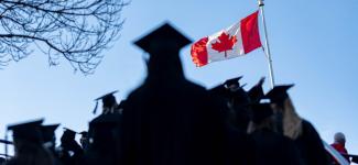 Canadian flag is raised against a blue sky background, with the shadow of a student in a grad cap in the foreground.
