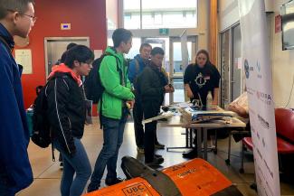 UBC IGEN graduate Darci May Nesbitt stands at a table while presenting information about UBC Aerodesign to a crowd of five people. 