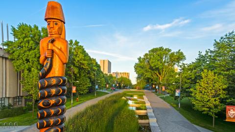 A tall totem pole featuring a human figure holding a serpant that wraps around them, stands at the entry to a long tree-lined boulevard.