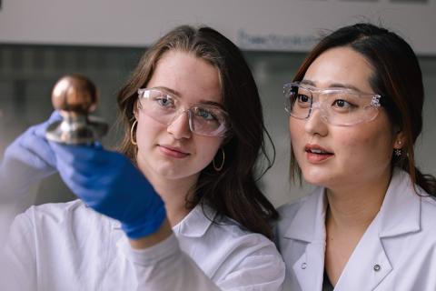 Two researchers stand side-by-side as they hold up and closely inspect a door handle encased in a copper coating