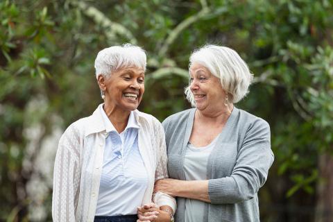 Two senior women conversing, walking together outdoors, with trees behind them. 