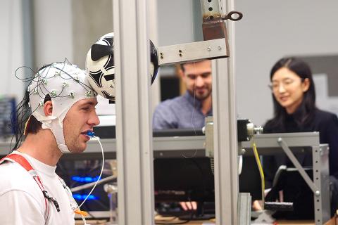 A study participant sits with EEG sensors on his head. In front of him is a mechanical arm with a soccer ball attached to simulate doing a mild header under controlled conditions.