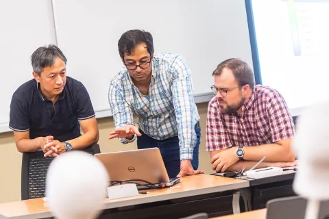 Three people look at a laptop, with white foam head forms in the foreground