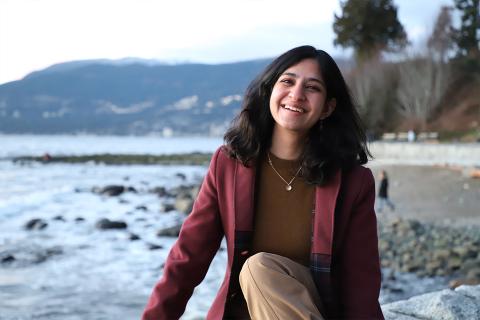 UBC Materials Engineering student Gauri Taneja smiles for the camera while seated at the beach with ocean and mountains in the distance.
