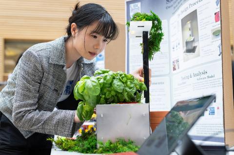 Student appears to be working on a project related to sustainable technology, possibly involving plant growth or environmental innovation, as they are examining a plant-growing setup, with research posters in the background explaining the design and analysis process