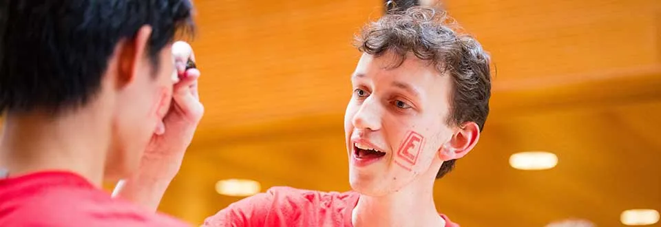 An engineering student with a temporary E tatoo on his face interacting with another student.
