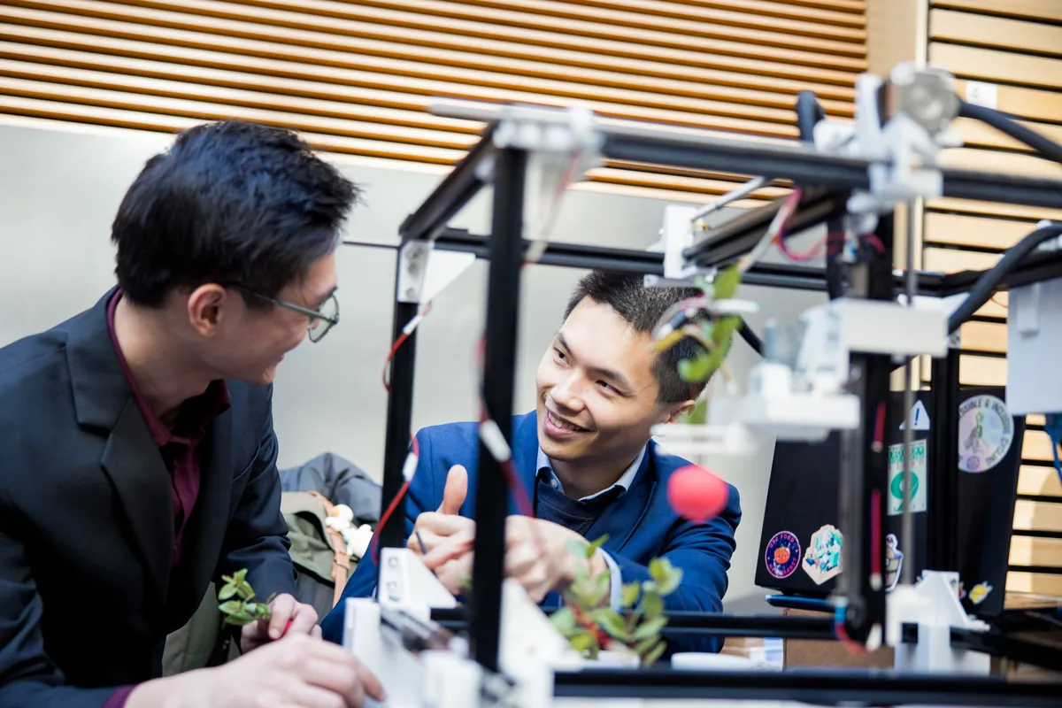 Two students discuss a small device transporting a radish with a robotic arm.