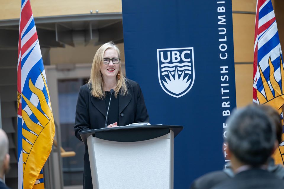 The Honourable Brenda Bailey speaks at a podium, in front of two BC flags and in front of a UBC banner