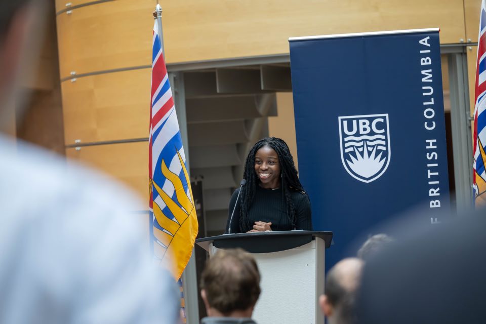 Woman student speaker speaks at a podium, flanked by BC flags and in front of a UBC banner